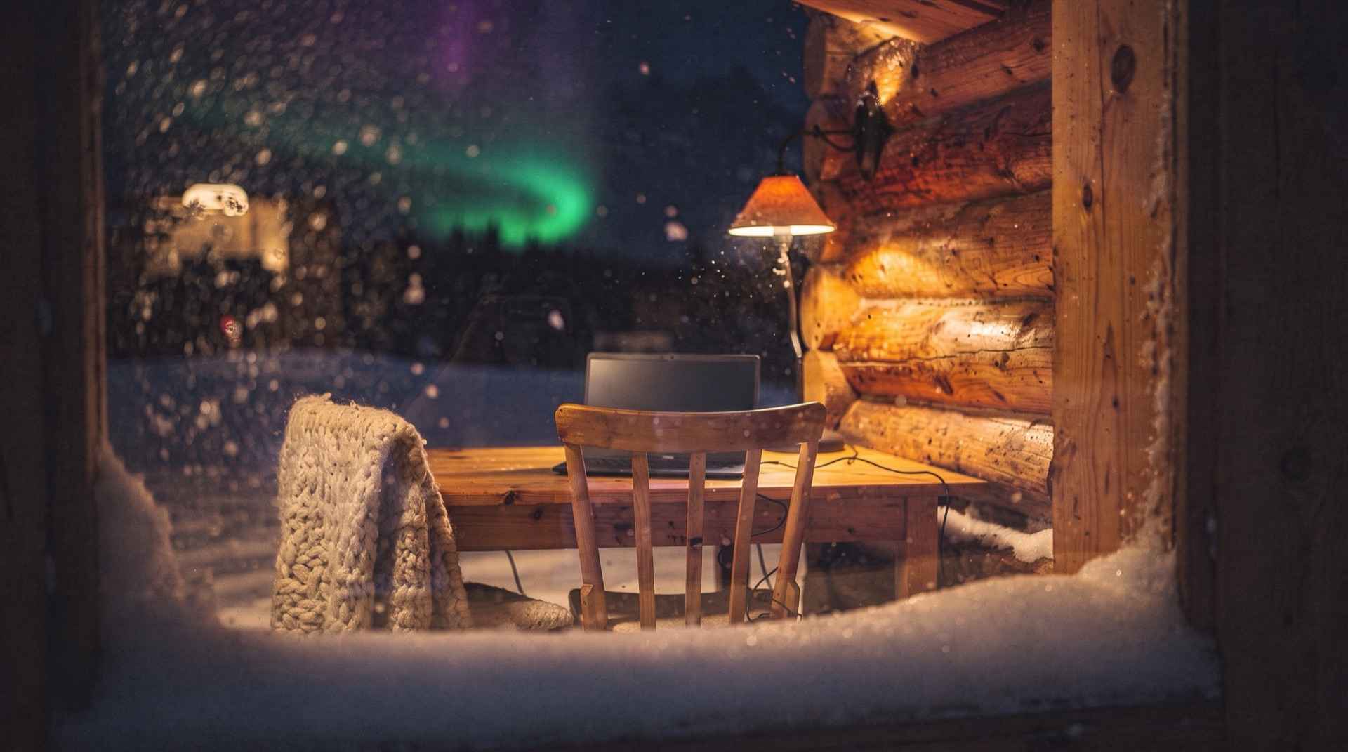 A warm cabin desk with a laptop seen through a snowy window