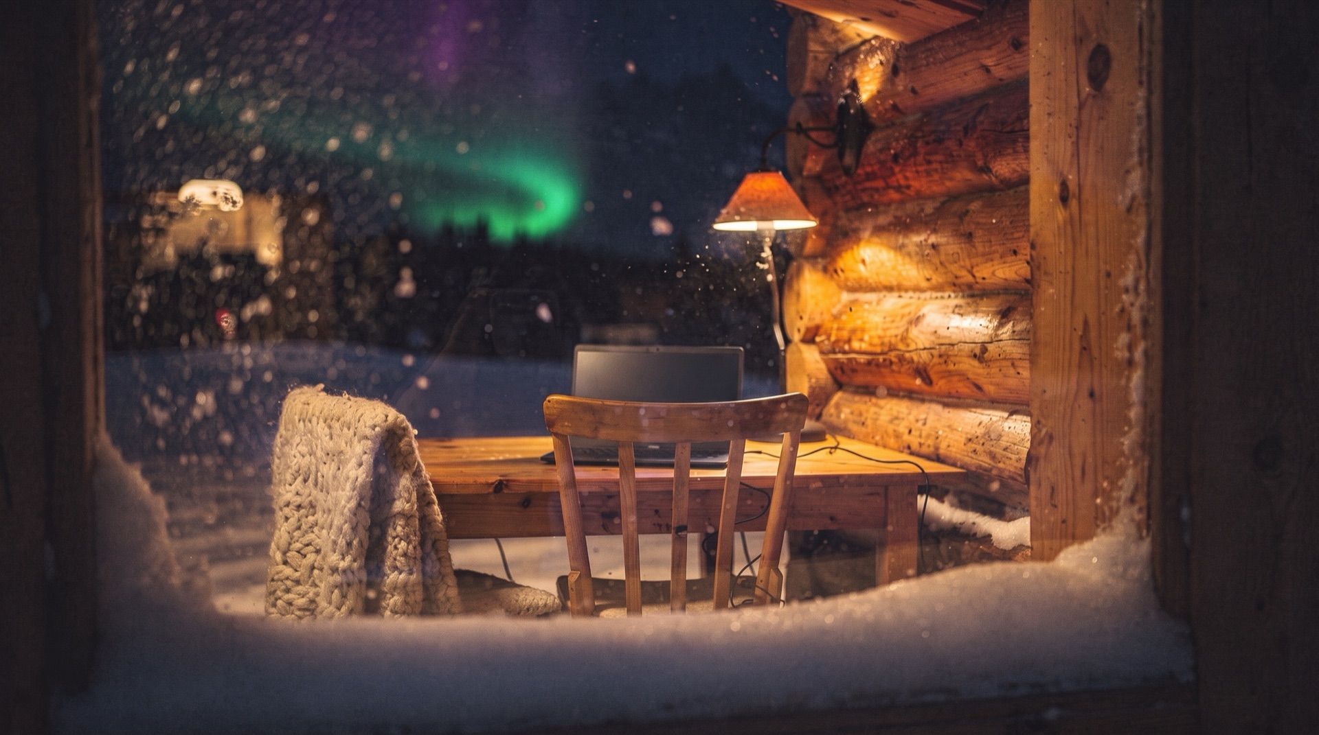 A warm cabin desk with a laptop seen through a snowy window